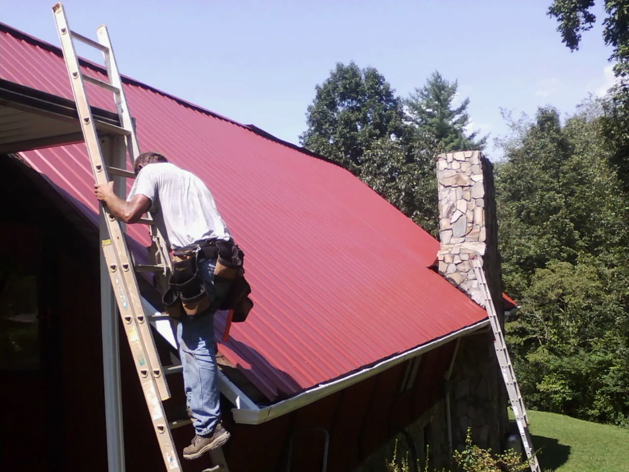 Roofer on a ladder beside a red metal roof and stone chimney — Soffit & Fascia Repair by Roofing Brothers in Rudd